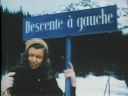En kvinna med toppluva under en skylt med texten "Descente à gauche", alptopp i bakgrunden.