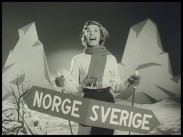 Alice Babs med halsduk och skidstavar bakom en skylt med texten NORGE / SVERIGE, fjällkuliss i bakgrunden.