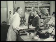 Två kvinnliga kunder handlar över disk av ett butiksbiträde i en gammaldags speceriaffär.
