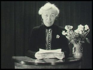 Kerstin Hesselgren med papper i hand vid talarbord med blomvas.