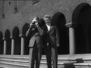 Två unga pojkar i kostymer framför Stockholms stadshus, en av pojkarna tar ett foto med en kamera.