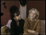 Två unga kvinnor i en soffa, en klänning hänger på väggen i bakgrunden.