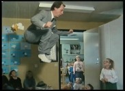 En vuxen man i kostym hoppar högt på en studsmatta inomhus omgiven av dagisbarn.