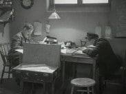 Två män sitter mittemot varandra vid varsitt skrivbord upptagna med pappersarbete.