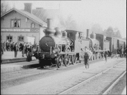 Tåg vid perrongen i Ludvika 1909. Människor runtomkring, stationshuset till vänster i bild. Järnvägsspår till höger i bild.