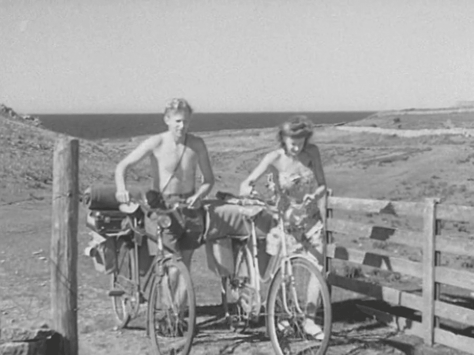Man och kvinna leder varsin fullpackad cykel genom grind nära havet.