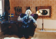 Åldrad man med medaljer på bröstet sitter i en soffa bredvid en svensk flagga som står på ett bord (leranimation).
