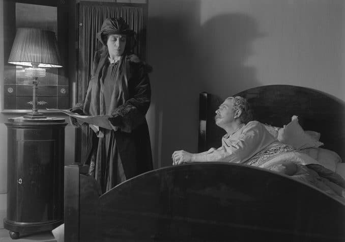 En man i en säng och en kvinna ståendes bredvid tittar på varandra.