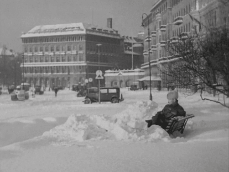 En man i pälsmössa sitter på en bänk i ett vintrigt Stockholm december 1931, bil och byggnader i bakgrunden.