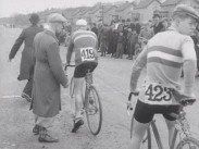 Två landsvägscyklister med nummerlappar och ett par funktionärer i långrockar i samband med tävlingsstart, publik i bakgrunden.