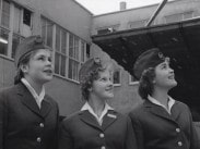 Tre unga kvinnor i uniform på rad blickar snett uppåt på någonting utanför bild.
