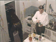 Två män står i ett kök nedstänkt med färg, färgburkar på bordet.