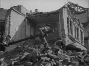 Två barn lyfter en träbjälke i rasmassorna framför ett sönderbombat hus.