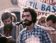 Skäggig man i blårutig skjorta talar i en mikrofon på en scen omgiven av människor, banderoller i bakgrunden.