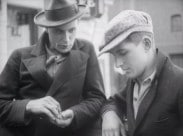 Två män med huvudbonader samtalar, den ene röker och den andre håller några mynt i handen.