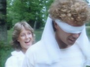 En man har en vit halsduk knuten över sin ögon, skrattande kvinna bakom honom.