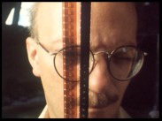 En man med mustasch och glasögon håller upp ett par smalfilmsremsor framför sitt ansikte.