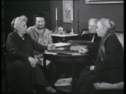 Fyra äldre kvinnor samlade kring ett bord, ett par ljusstakar i bakgrunden.