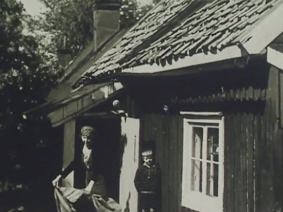 Kvinna och pojke framför gammal träkåk.