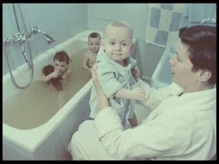 Kvinna håller spädbarn i morgonrock. Två småpojkar badar badkar i bakgrunden.