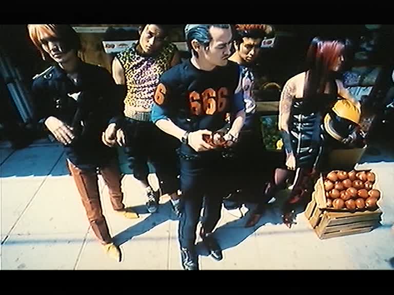 Fem ungdomar i trendiga kläder står på gatan utanför en fruktaffär.