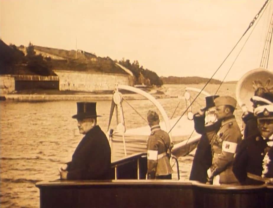 Finska republikens president Relander i hög hatt på båt på väg in till Stockholm 1925, militärer ombord bakom honom.