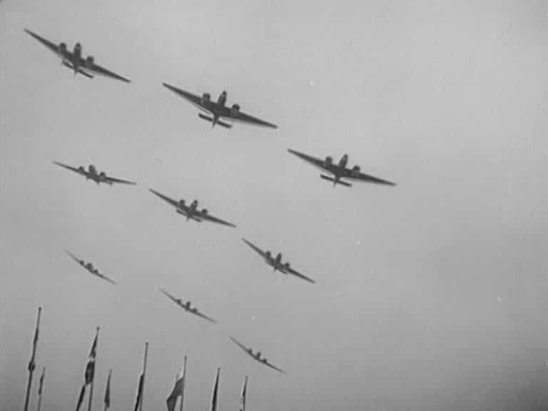 Nio militärflygplan i flygformation, några flaggstänger längst ner i bild.
