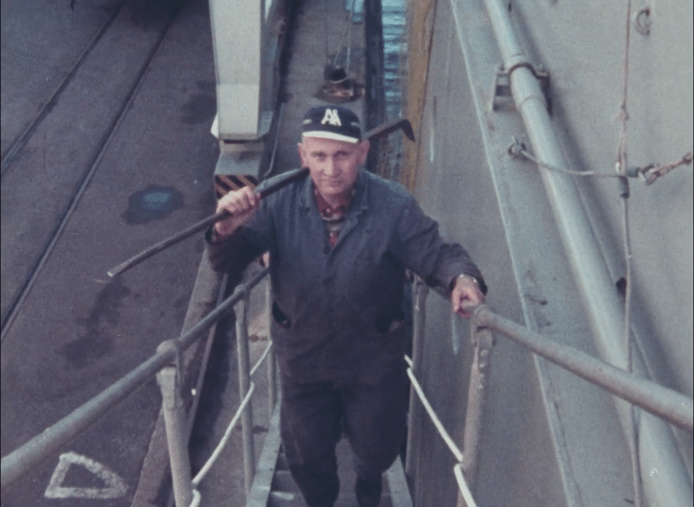 Hamnarbetare iklädd arbetsoverall i smal trappa med verktyg på axeln.