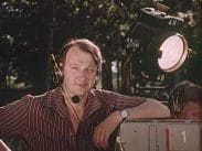 En man med inspelningshörlurar med mikrofon som står utomhus med ena armbågen lutad mot en filmkamera, en filmlampa i högre övre hörnet.