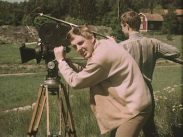 Lars Amble bakom en filmkamera i naturen, bakom honom en man med ryggen mot kameran som tar upp ljud med en mikrofonbom.