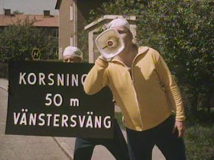 Man i gul träningsoverallsjacka håller en megafon framför sitt ansikte. Ytterligare en man till vänster i bild håller en skylt med texten "Korsning 50 m vänstersväng".