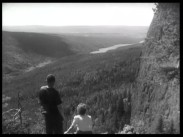 En man och en kvinna blickar ut över ett storslaget skogslandskap med en ringlande älv, bergsklippa till höger i bild.