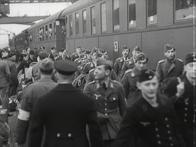 Uniformsklädda soldater på perrong. Tåg i bakgrunden, några soldater hänger ut ur fönstren.