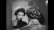 Kvinna med ryggen mot kameran med sänkt blick framför spegel, hennes ansikte syns däri.
