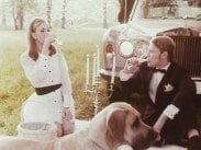 Ett par dricker champagne utomhus på en picknickfilt med en kandelaber framför en parkerad Rolls Royce, hund i förgrunden.