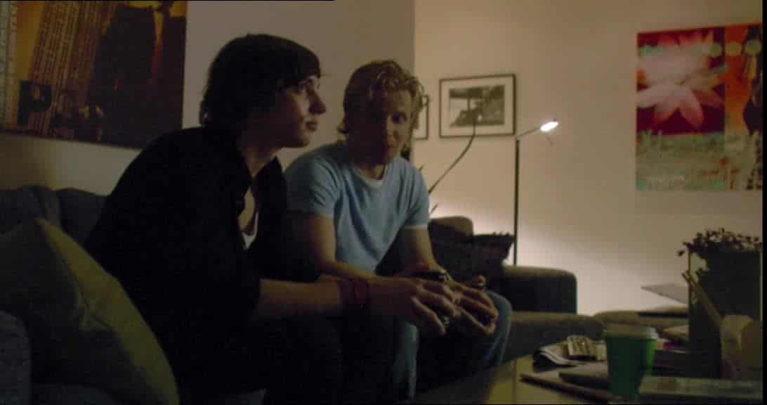 Två unga män sitter i en soffa i ett vardagsrum, dämpad belysning.