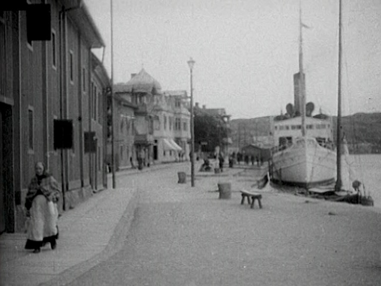 Ett par båtar ligger förtöjda längs kajen i Marstrand. En gumma i förkläde går på trottoaren.