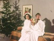 En man och en kvinna sitter i samma julklappssäck, mannen håller upp några chokladkakor - julgranar i bakgrunden.