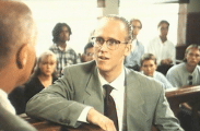 En ung åklagare i kostym och med glasögon i en rättssal, ett förhörsvittne fotad bakifrån i förgrunden och åhörare i bakgrunden.