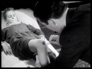 En pojke ligger på en sjukhusbrits och får ett sår på ena knät omhändertaget av en man i mörk kostym.