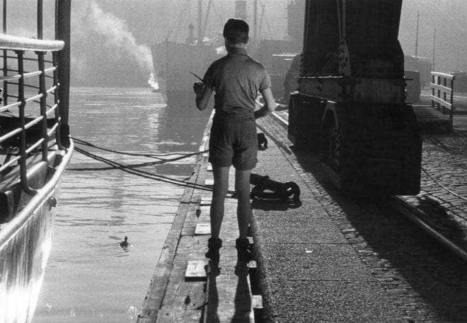 Pojke med ryggen mot kameran ståendes på kaj, fartyg i bakgrunden.