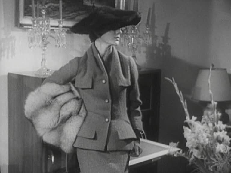 Mannekäng iklädd dräkt, hatt och med en päls under armen visar upp Diors höstmode 1954.
