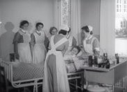 Patient i sjukhussäng omgiven av sex sköterskor.