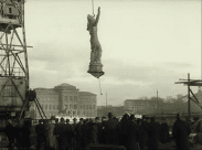 Staty hängande i luften med krok och kedja upptill, lyftkran till vänster i bild och Nationalmuseet i Stockholm i fonden.