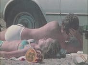 Ett par i badkläder som solar på en sandstrand. En plasthink i förgrunden och en vit bil i bakgrunden.