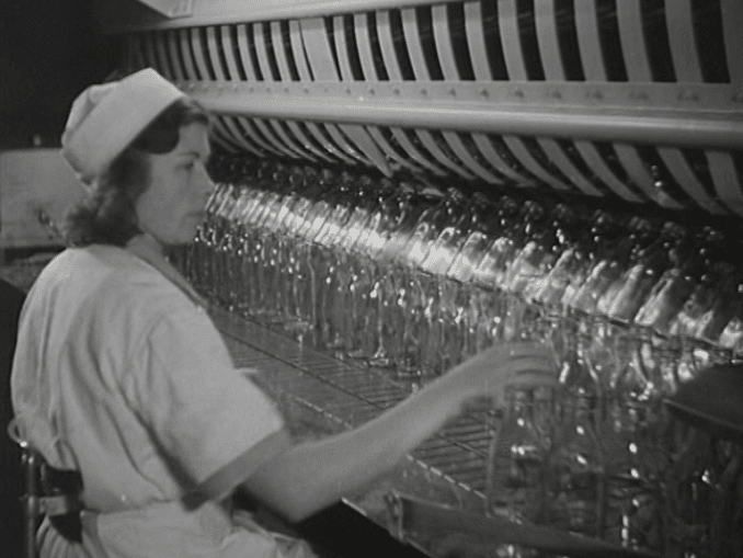 En kvinna i arbetskläder vid ett löpande band fyllt av tomma mjölkflaskor i glas.
