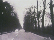 Foto genom vindrutan på bil, back spegel i nedre högra hörnet. Bilar på landsväg kantad av träd på båda sidor.