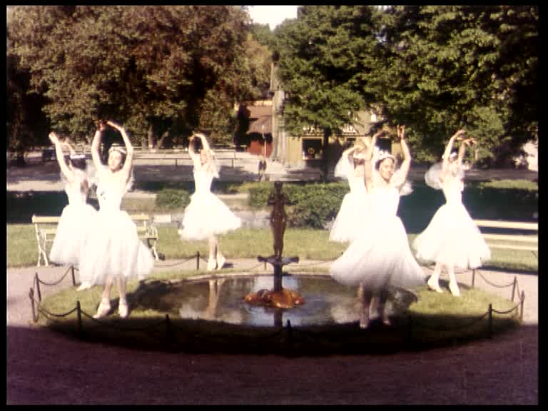 Sex kvinnor i vita klänningar dansar balett kring en utomhusfontän.