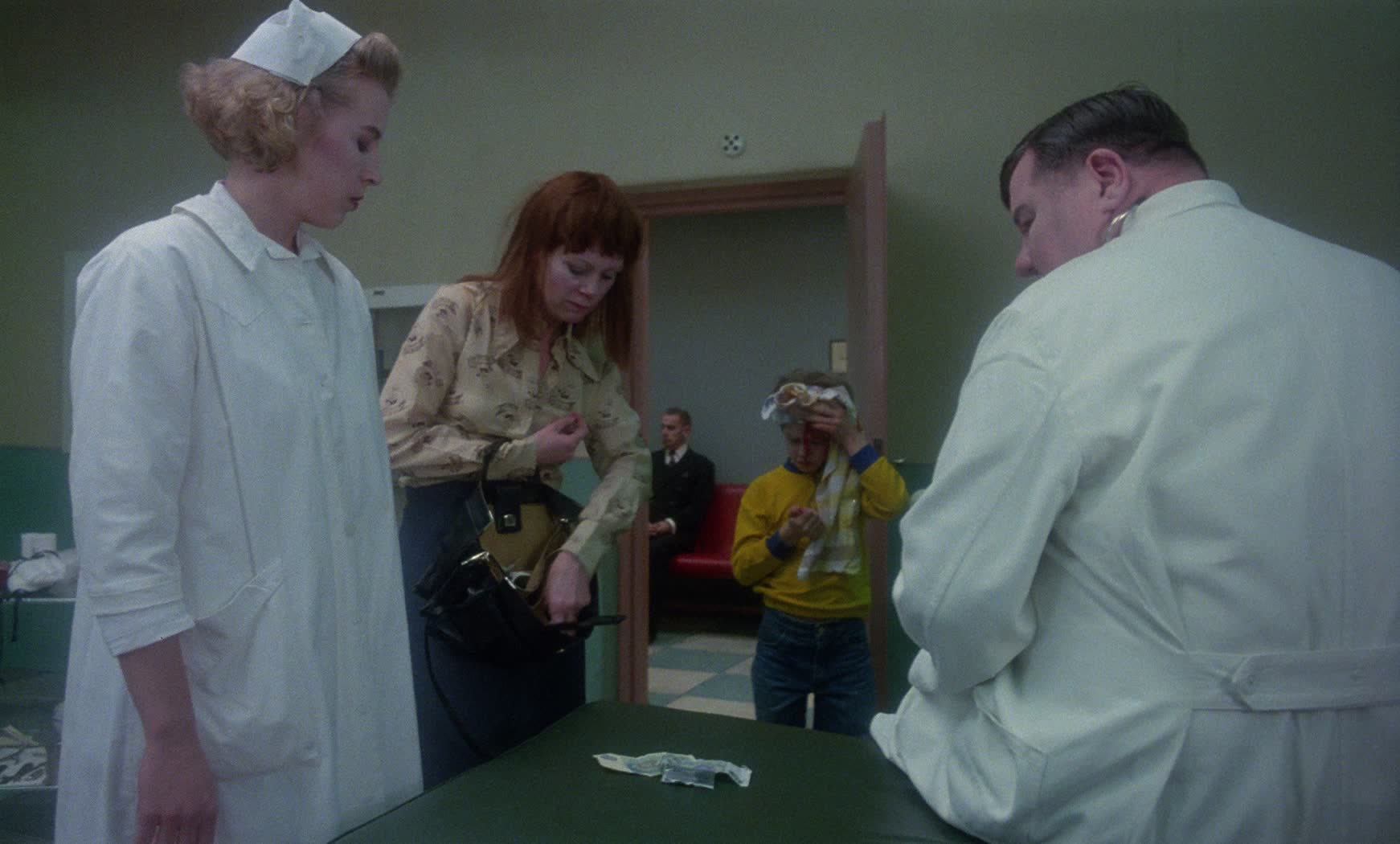 En mamma på besök på en läkarmottagning tillsamman med sin skadade son har lagt upp sedlar på en brits och letar efter fler i sin handväska noga övervakade av en läkare och en sjuksköterska.