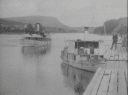 Båt i trafik på Ångermanälven. Ytterligare en båt ligger vid en brygga till höger i bild. På bryggan står en man i kostym.